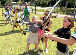 Taking aim at opportunity: A Mercer lawyer’s mission to empower youth through archery Children and adults practice archery outdoors on a grassy field, aiming bows at targets in the distance.