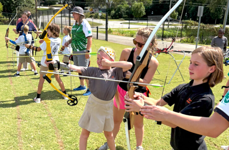 Children and adults practice archery outdoors on a grassy field, aiming bows at targets in the distance.