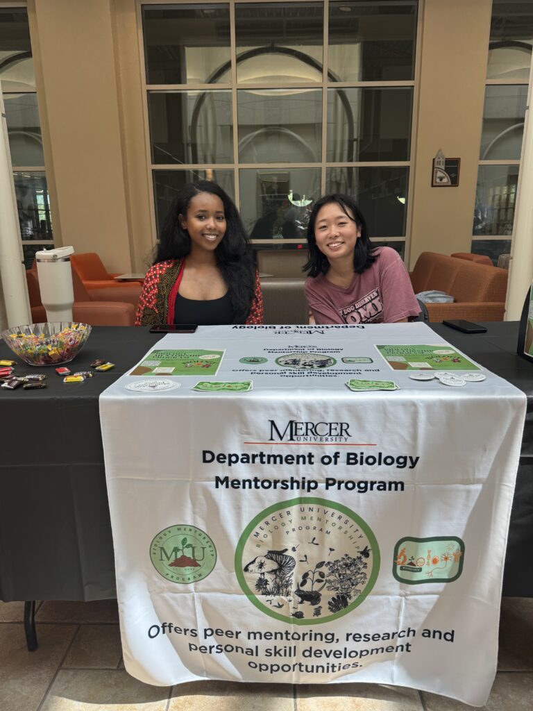 Two people sit at a table promoting the Mercer University Department of Biology Mentorship Program in an indoor space.