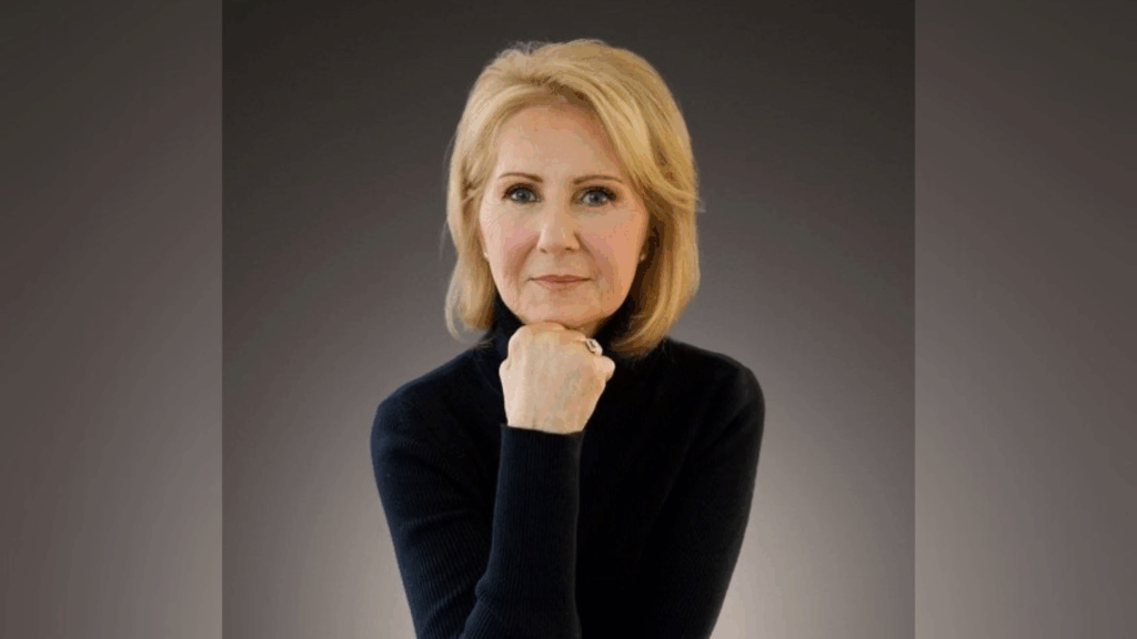 A woman with blond hair wearing a black turtleneck poses with her chin resting on her hand against a dark background.