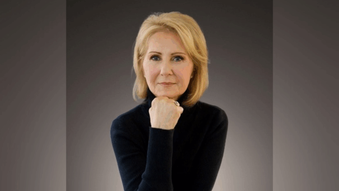 A woman with blond hair wearing a black turtleneck poses with her chin resting on her hand against a dark background.