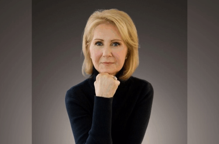 A woman with blond hair wearing a black turtleneck poses with her chin resting on her hand against a dark background.