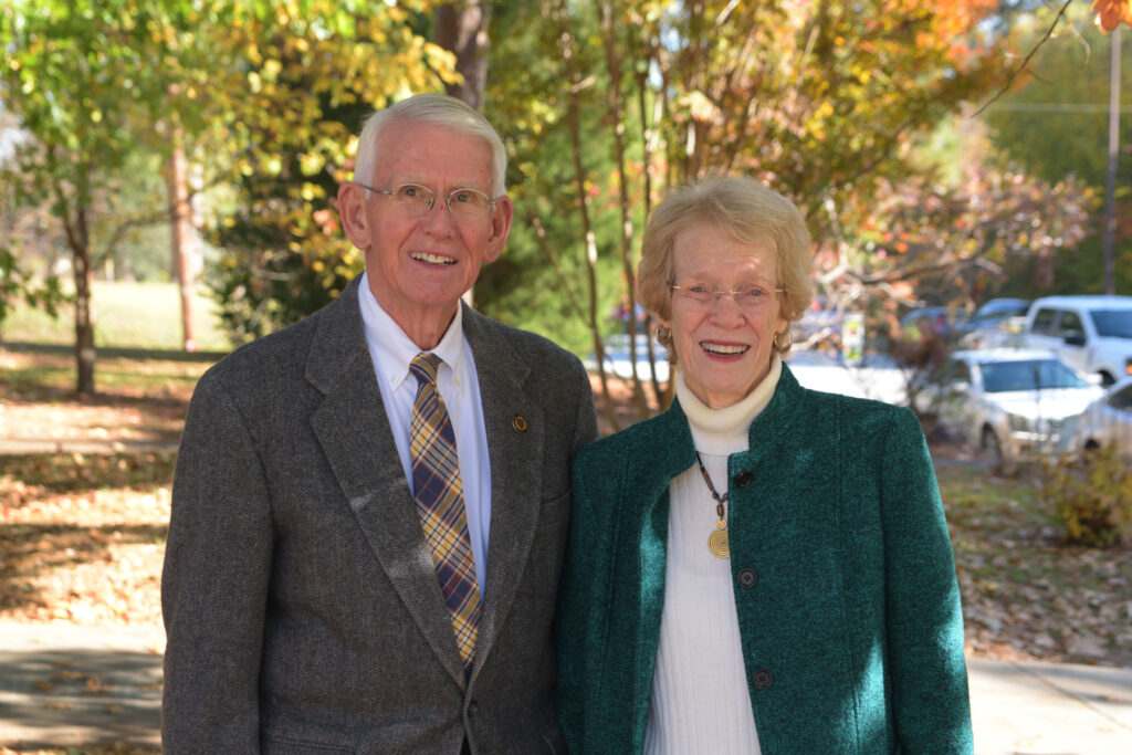 An elderly man and woman stand outside on a sunny day, smiling at the camera with trees and parked cars behind them.