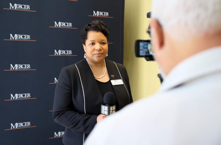 A woman is interviewed on camera in front of a Mercer University backdrop, holding a microphone.