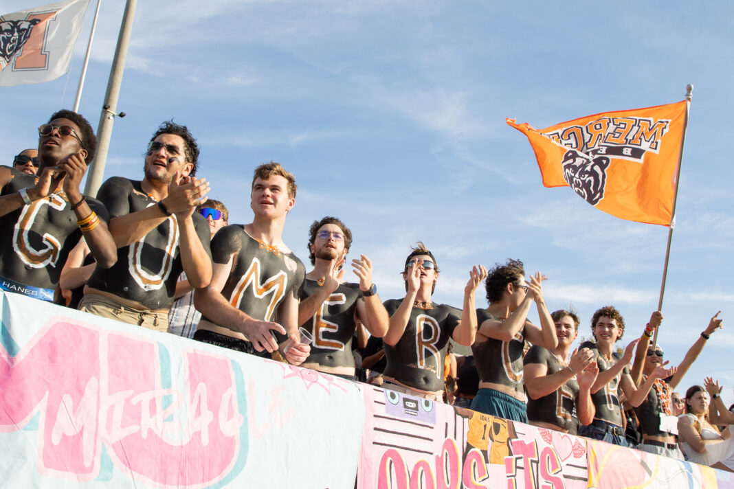 A group of fans with painted chests stands in a row cheering, while one holds an orange flag with a bear logo.