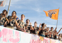 3 things to know about Mercer’s playoff game vs. South Dakota: Dec. 6, 2025 A group of fans with painted chests stands in a row cheering, while one holds an orange flag with a bear logo.