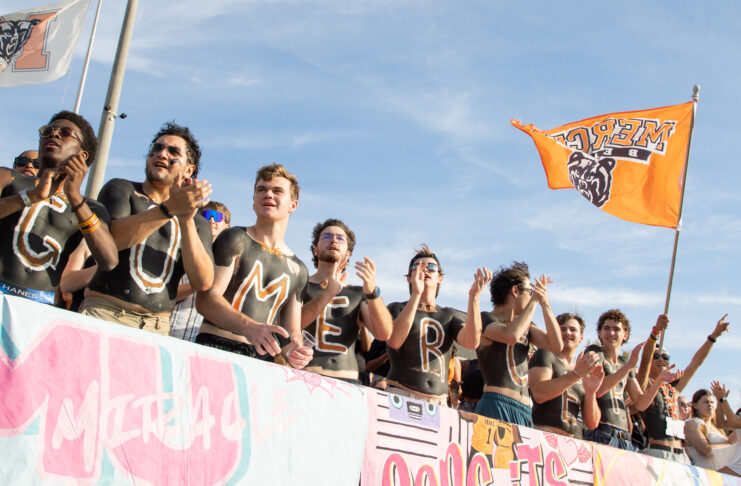 A group of fans with painted chests stands in a row cheering, while one holds an orange flag with a bear logo.