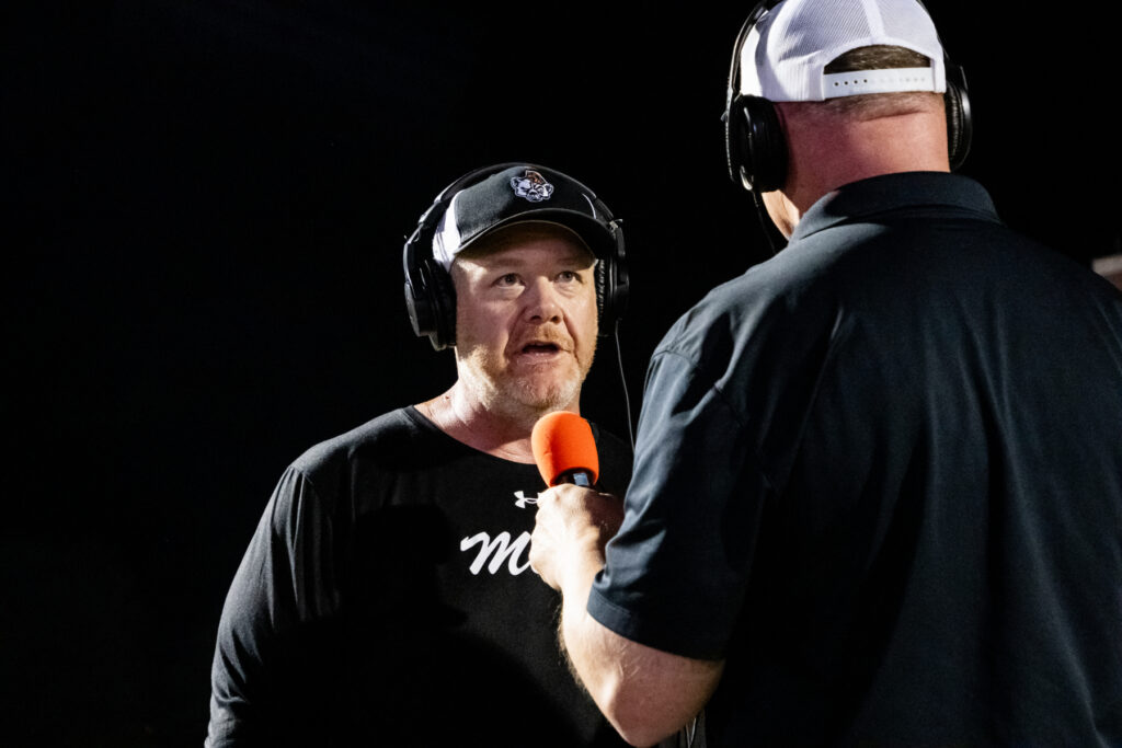 Two men wearing headsets talk during an interview; one holds an orange microphone toward the other.