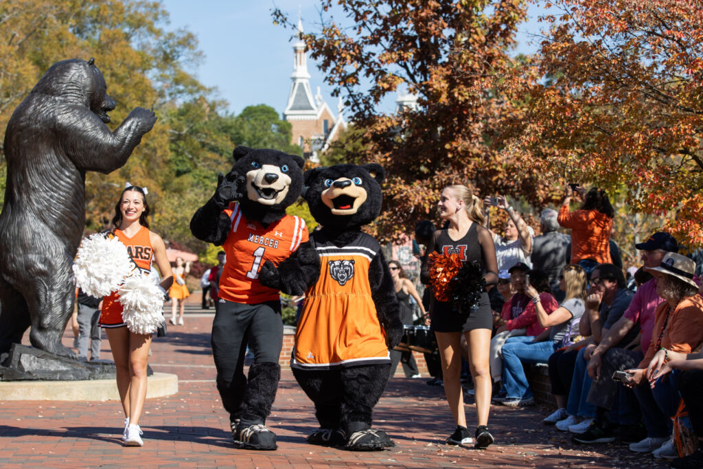 Two bear mascots and cheerleaders walk near a bear statue as people watch during a college event on a sunny day.