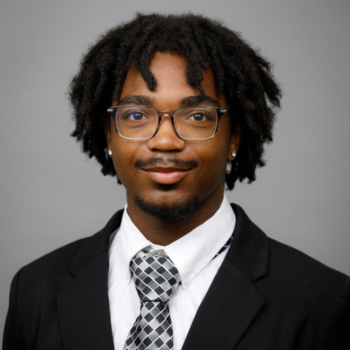 Man with short dreadlocks, glasses, and a goatee wearing a suit and patterned tie poses against a plain gray background.