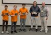 Binary Bears place first in regional programming competition Four young men in orange shirts hold plaques next to an older man in business casual attire, posing indoors.