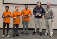 Binary Bears place first in regional programming competition Four young men in orange shirts hold plaques next to an older man in business casual attire, posing indoors.