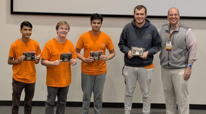 Binary Bears place first in regional programming competition Four young men in orange shirts hold plaques next to an older man in business casual attire, posing indoors.