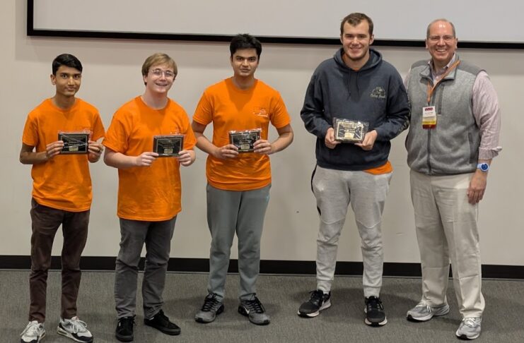 Four young men in orange shirts hold plaques next to an older man in business casual attire, posing indoors.