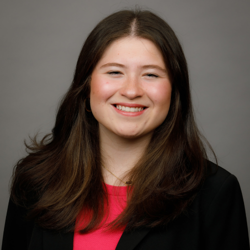 A young woman with long brown hair smiles, wearing a black blazer and pink top, in front of a plain gray background.