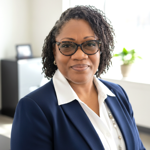 Woman in a navy blazer and glasses stands in a bright office, smiling slightly at the camera.