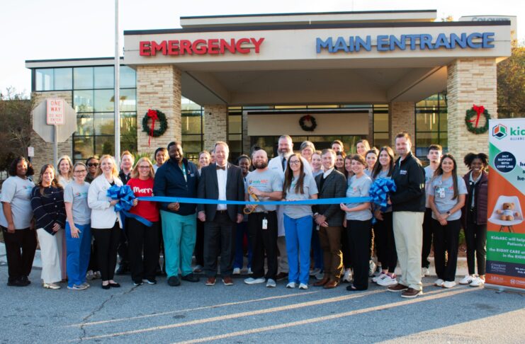 A group of people stands outside a hospital entrance, holding a large blue ribbon for a ribbon-cutting ceremony.
