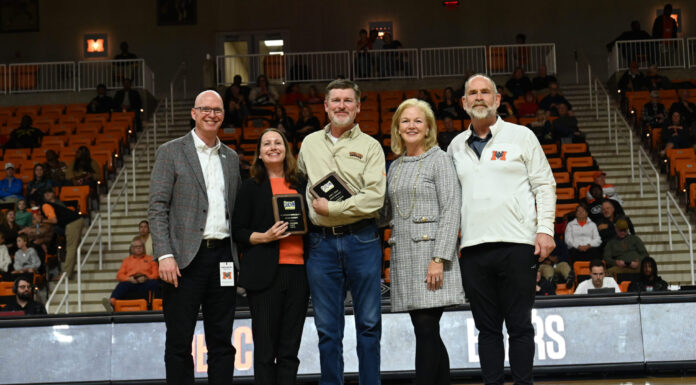 Cardelino, Steele honored on All-Southern Conference Faculty and Staff Team Five people stand on a basketball court during a ceremony, with one person holding a plaque and spectators in the background.