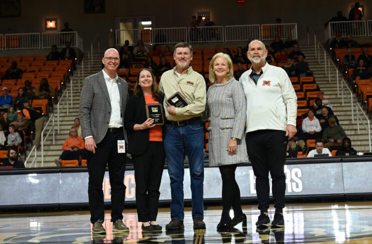 Five people stand on a basketball court during a ceremony, with one person holding a plaque and spectators in the background.