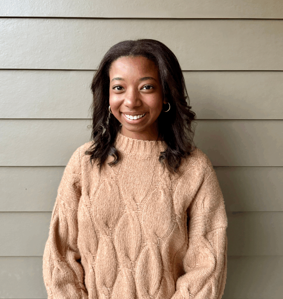 A woman in a tan cable-knit sweater stands in front of a beige paneled wall, smiling at the camera.