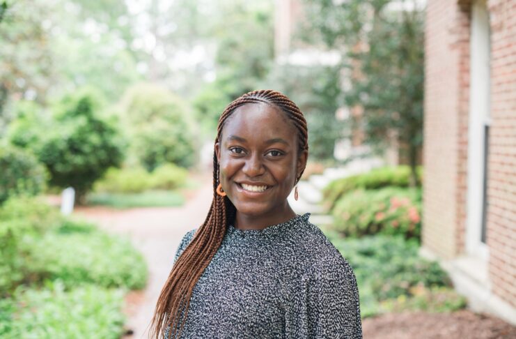 Young woman with braided hair smiles outdoors, standing near a brick building with greenery in the background.