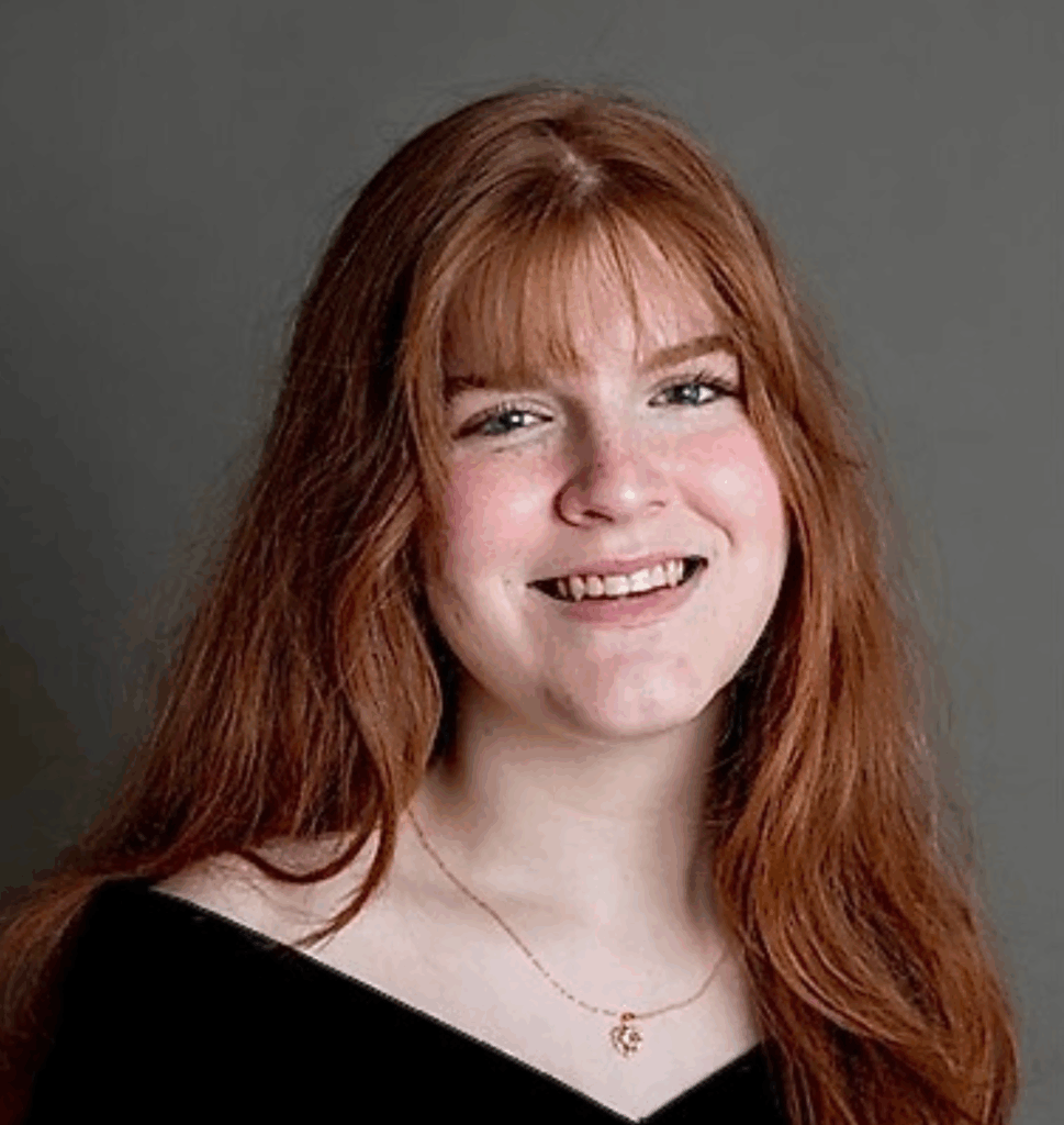 Young woman with long red hair, wearing a black top and gold necklace, smiling in front of a gray background.