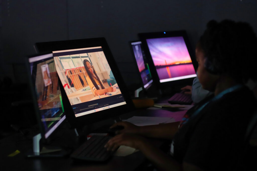A person at a computer edits a video in a dimly lit room with multiple monitors displaying various images.