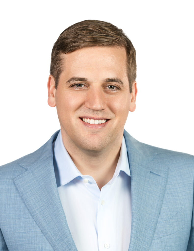 A man with short brown hair wearing a light blue suit jacket and white shirt, smiling at the camera.