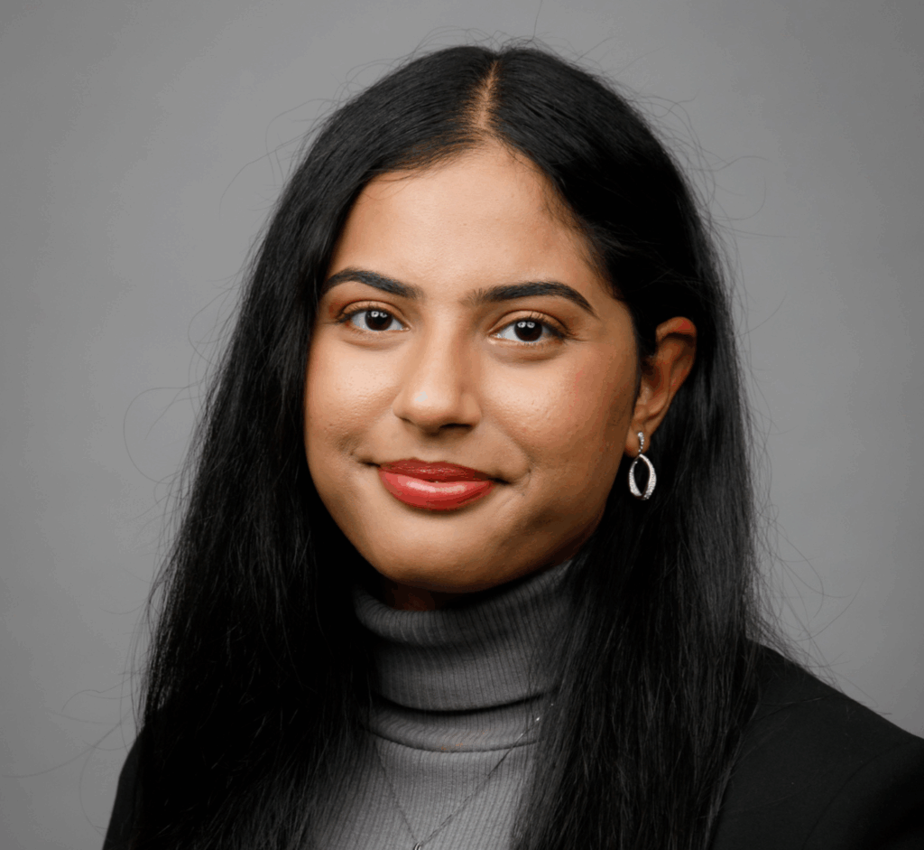 A woman with long black hair, wearing a gray turtleneck and black jacket, poses in front of a gray background.