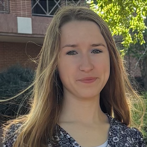 A young woman with long brown hair stands outside in sunlight, wearing a black and white patterned top.