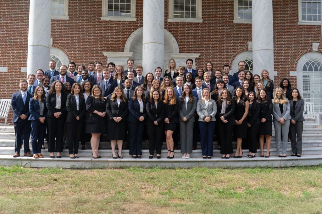 A large group of professionally dressed people pose for a photo on outdoor steps in front of a brick building with columns.