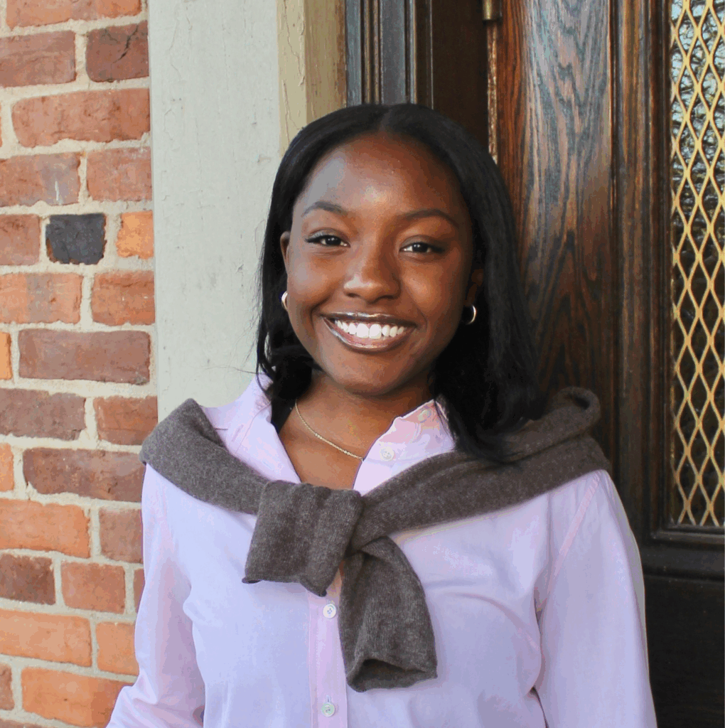 A woman in a pink shirt with a brown sweater draped over her shoulders stands smiling in front of a brick wall and doorway.