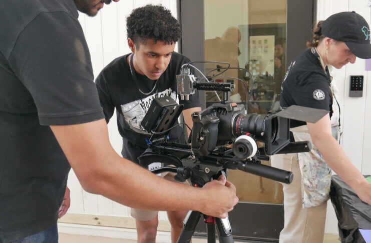 Three people set up and adjust a professional video camera mounted on a tripod indoors.