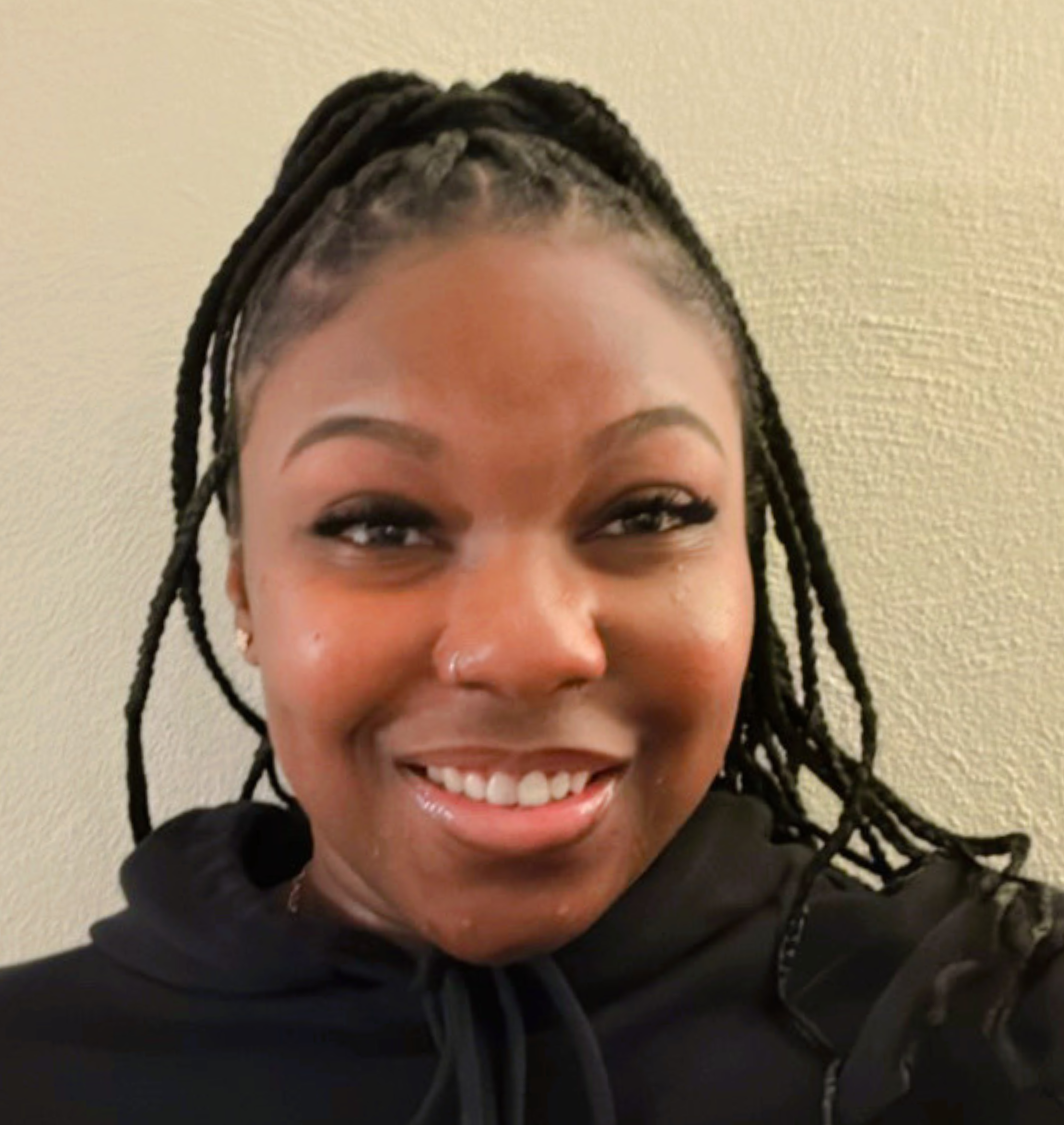 A woman with braids, wearing a black hoodie, smiles at the camera against a plain beige wall.