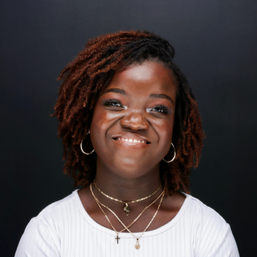 Woman with short curly hair, wearing a white top and layered necklaces, smiles against a plain dark background.
