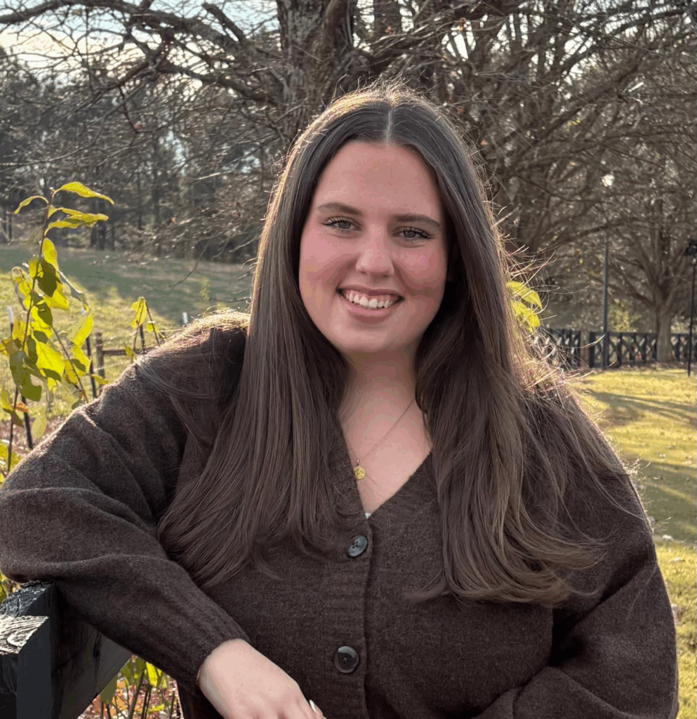 A woman with long brown hair wearing a brown sweater smiles outdoors near a wooden fence and trees.