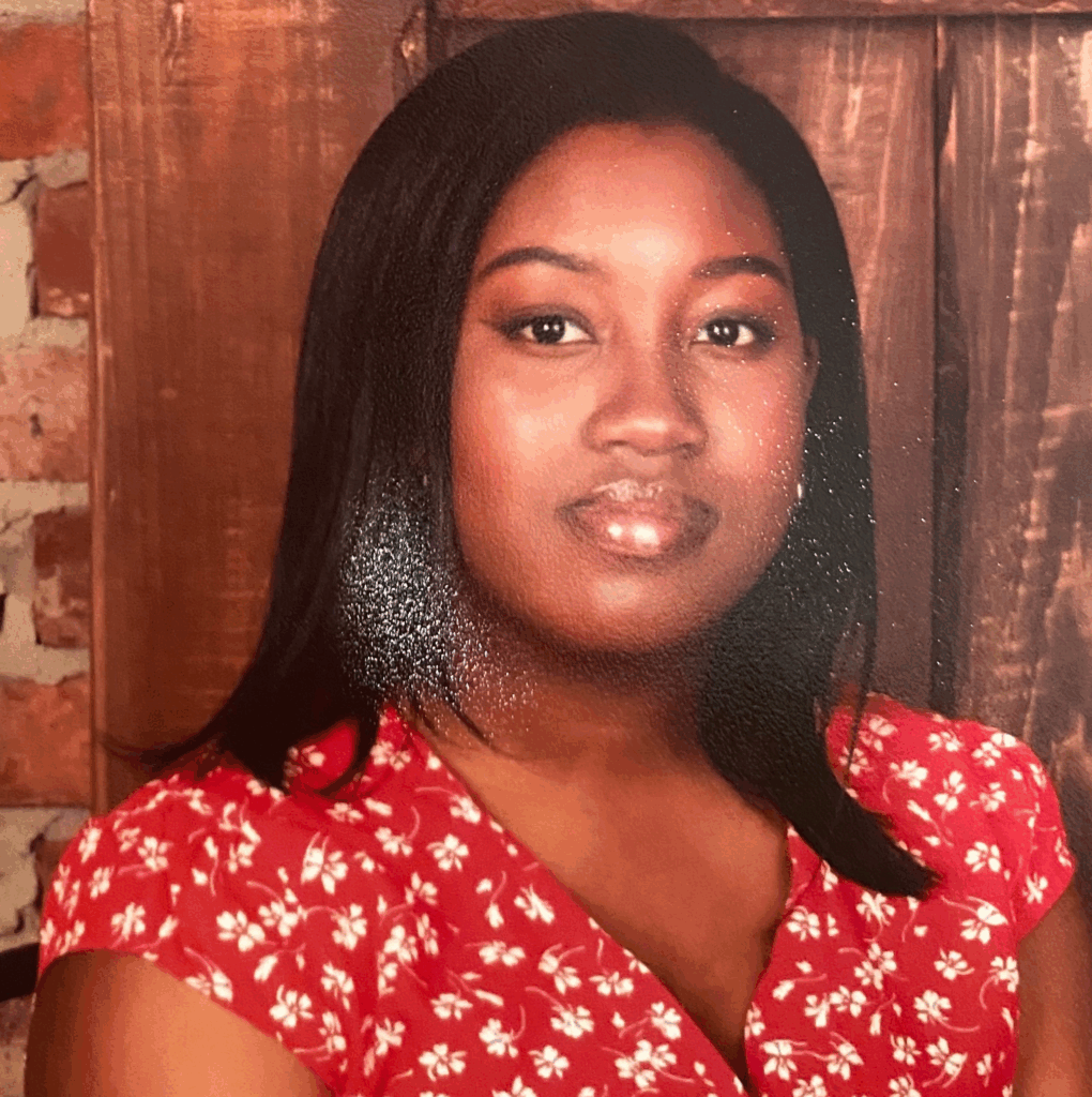 Woman with straight black hair wearing a red floral top, seated in front of a wooden and brick background.