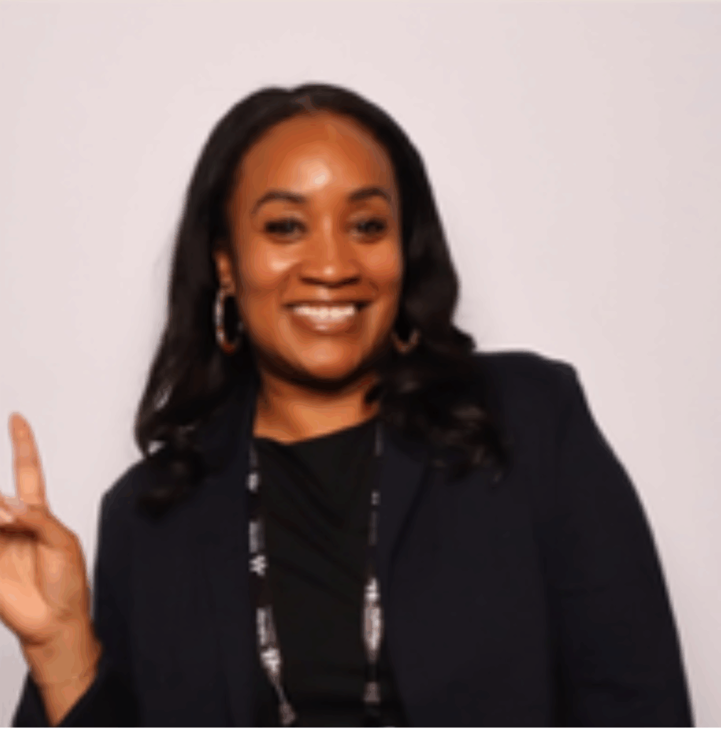 Woman with long dark hair smiles at the camera, wearing a dark blazer and holding up her left hand with one finger raised.