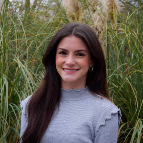 Woman with long brown hair wearing a gray top, smiling in front of tall green grass outdoors.