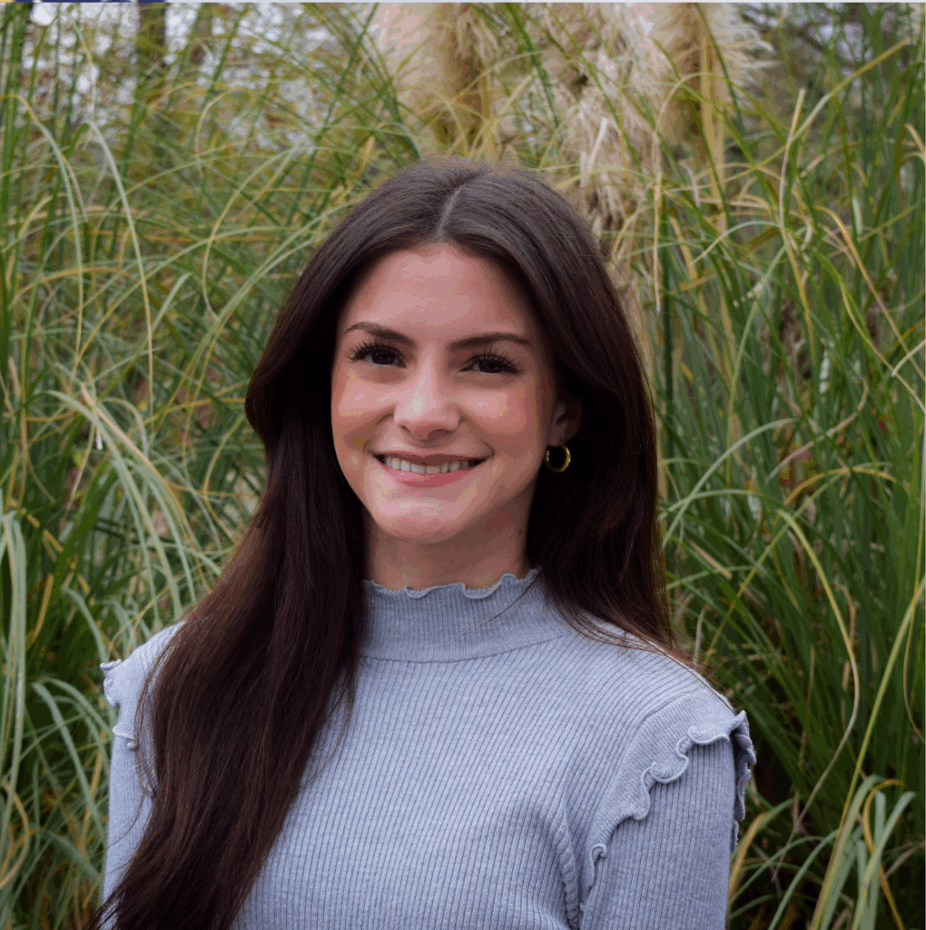 Woman with long brown hair in a gray top stands outdoors in front of tall green grass, smiling at the camera.