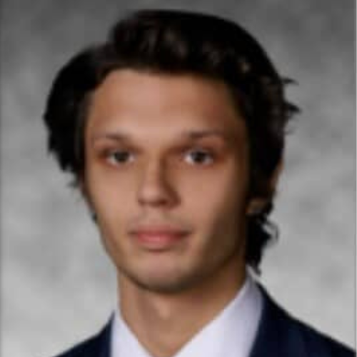 A young man with medium-length dark hair wearing a suit and tie poses against a plain, gray background.