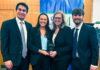 Mercer Law advocacy teams close out successful competition season Four people in business attire smile and hold an award plaque in front of a blue and green banner indoors.