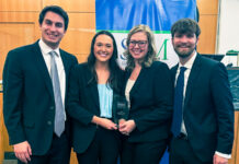 Mercer Law advocacy teams close out successful competition season Four people in business attire smile and hold an award plaque in front of a blue and green banner indoors.