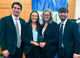 Mercer Law advocacy teams close out successful competition season Four people in business attire smile and hold an award plaque in front of a blue and green banner indoors.