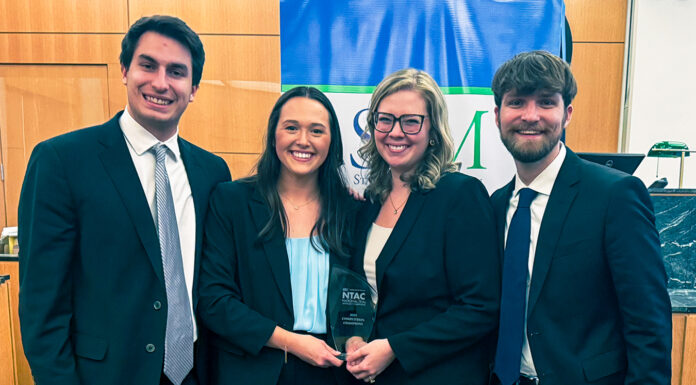 Mercer Law advocacy teams close out successful competition season Four people in business attire smile and hold an award plaque in front of a blue and green banner indoors.