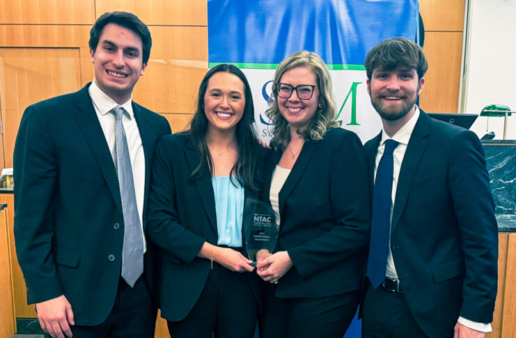 Four people in business attire smile and hold an award plaque in front of a blue and green banner indoors.
