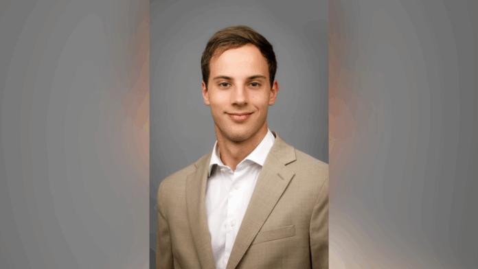 working file A young man in a beige suit jacket and white shirt poses against a plain gray background.