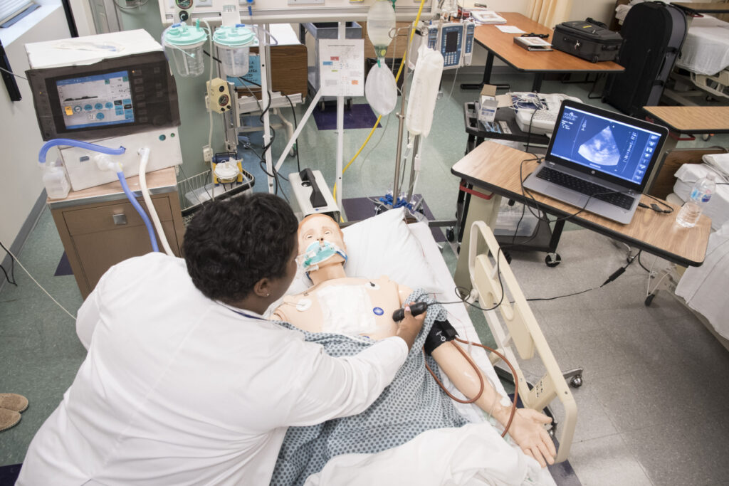 A medical professional examines a medical training mannequin in a hospital room equipped with monitors and medical devices.