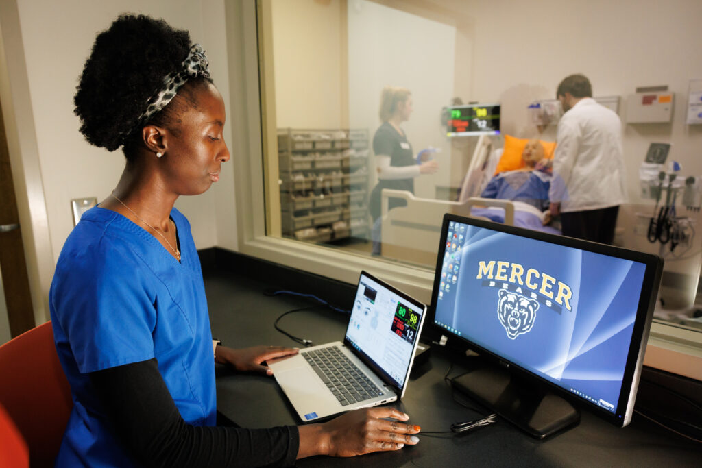 A person in scrubs monitors a laptop and desktop, while nursing students attend to a high-fidelity mannequin in a hospital room behind glass.