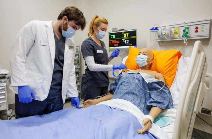 Two Mercer nursing students practice patient care on a medical mannequin in a hospital room with monitoring equipment.